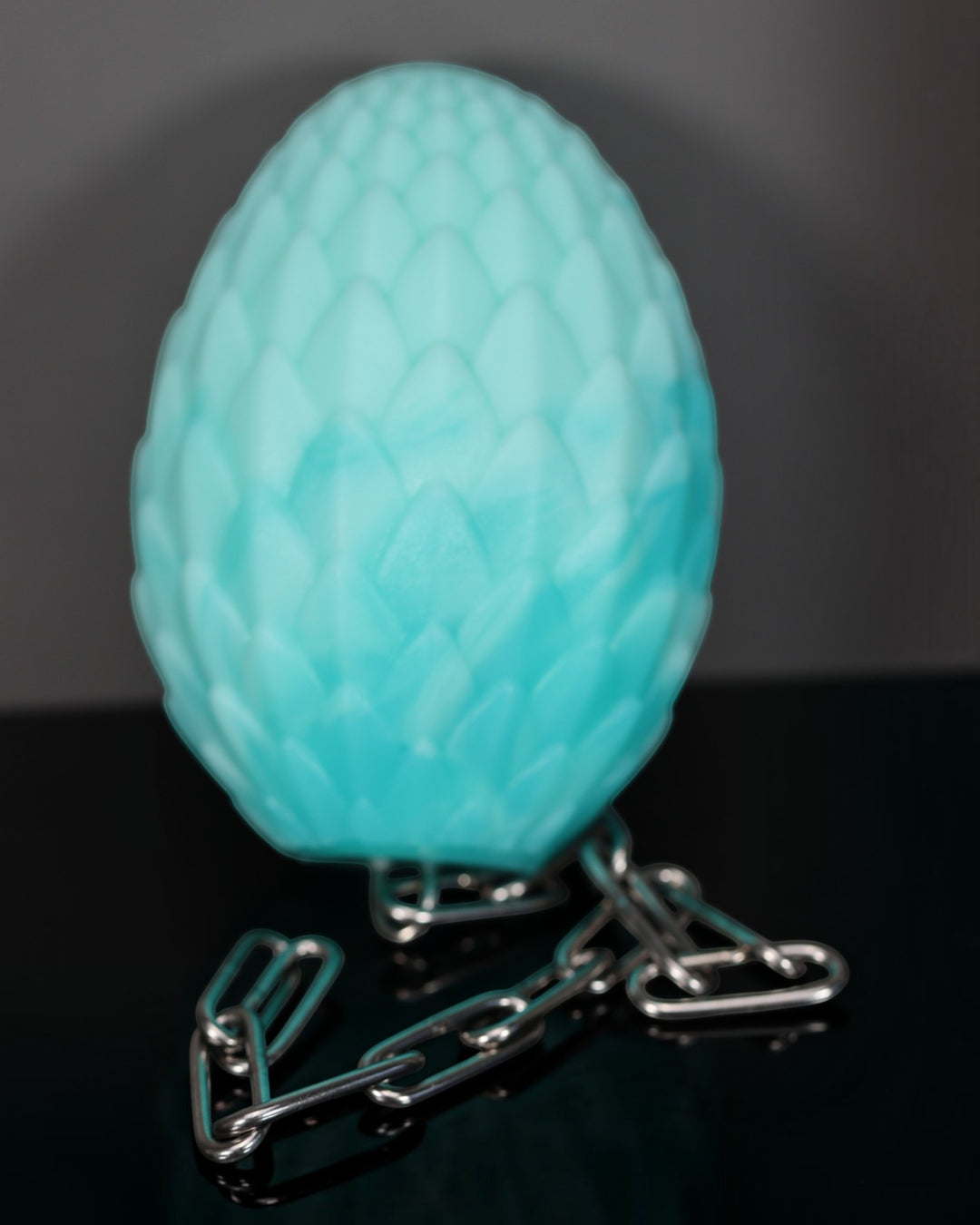 A textured, egg-shaped turquoise blue light or lampshade-like object displayed upright on a reflective black surface with a dark gray background, supported by a short section of silver metal chain.